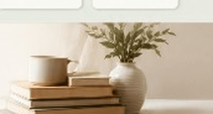 Livros empilhados com caneca e planta sobre superfície clara.